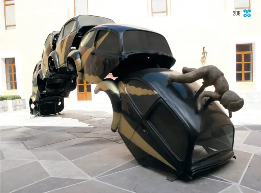 Fotografía cercana de la obra *Sobreponiéndose* (2008): carrocerías de vochitos negros y dorados que forman un arco, con una figura humana de barro trepando sobre la pieza superior, en un patio con piso de piedra.