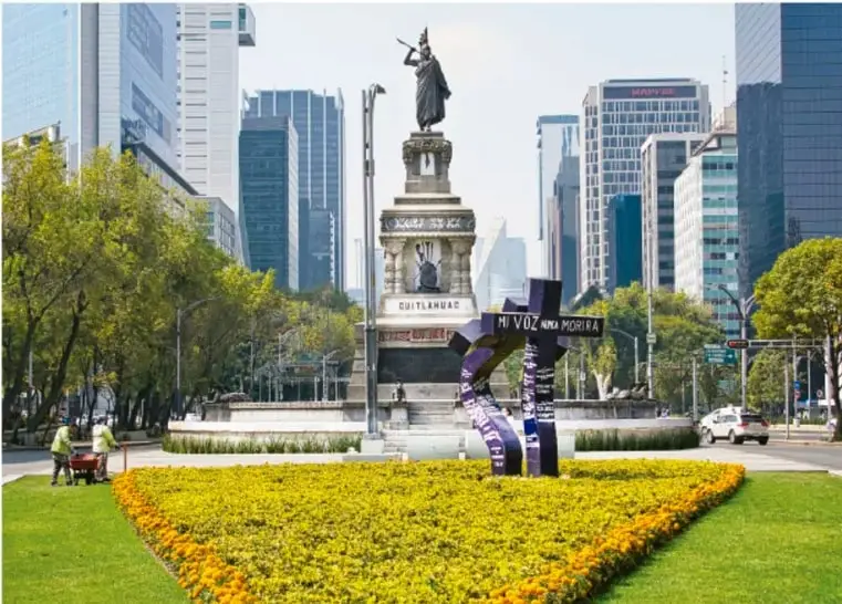 Fotografía de una avenida urbana en la Ciudad de México: glorieta con la estatua de la colona Cuitláhuac, jardín de flores amarillas en primer plano, edificios de oficinas al fondo y una estructura artística con cintas moradas que dice 'Mi voz una historia'.