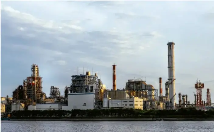 Fotografía de una gran planta industrial o petroquímica a orillas de un cuerpo de agua, con chimeneas y estructuras metálicas.