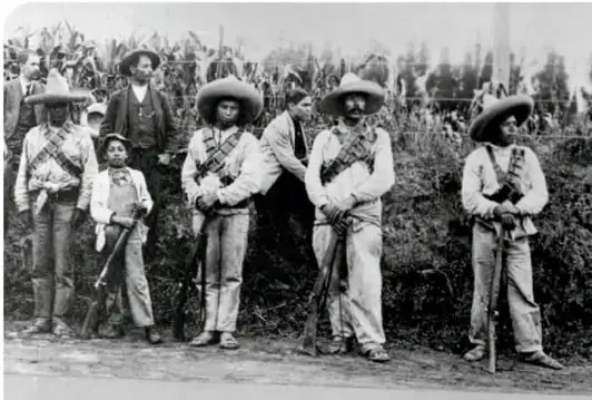 Fotografía en blanco y negro de un grupo de zapatistas en contexto agrícola, con sombreros y armas.