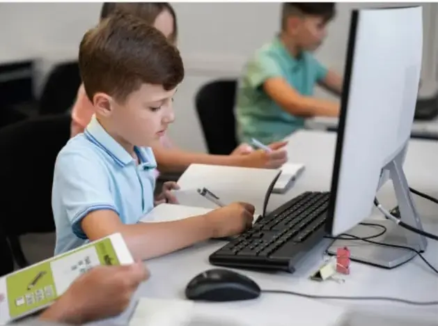 Niños trabajando en computadoras en un aula escolar, uno de ellos escribe en un cuaderno junto a un teclado.