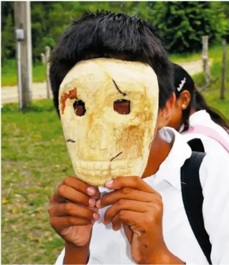 Niño de camisa blanca que sostiene ante su rostro una máscara tallada en madera clara con ojos y boca abiertos; al fondo pasto y cielo.