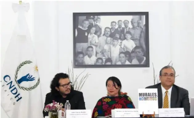 Fotografía de una conferencia de prensa de la CNDH. Aparecen tres personas sentadas ante una mesa, con una bandera de la CNDH a la izquierda y un cuadro con una fotografía grupal al fondo. Sobre la mesa hay un cartel que dice 'MURALES POR NUESTROS DERECHOS'.