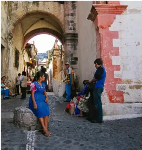 Fotografía de una calle empedrada con arcos coloniales y personas de la comunidad, incluidas mujeres con trajes tradicionales.