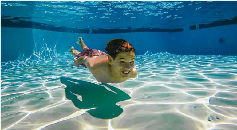 Fotografía de un niño nadando bajo el agua en una piscina, con su sombra proyectada en el fondo.