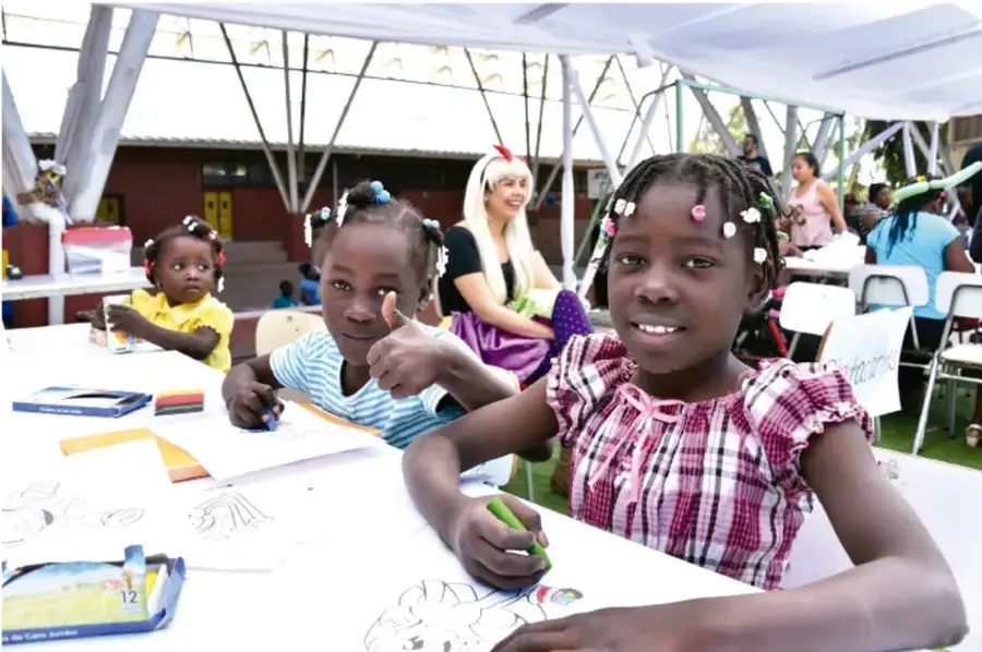 Fotografía de niñas y niños de distintas procedencias dibujando y conviviendo en una mesa bajo una carpa, acompañados por una docente; representa la interculturalidad y la inclusión en la escuela.