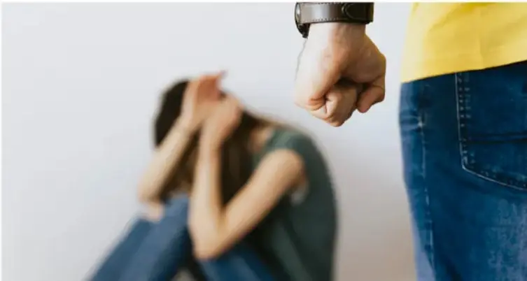 Fotografía de una persona adulta con el puño cerrado y actitud amenazante frente a una niña sentada en el suelo, encogida y cubriéndose la cara con las manos. Representa una situación de violencia o maltrato.