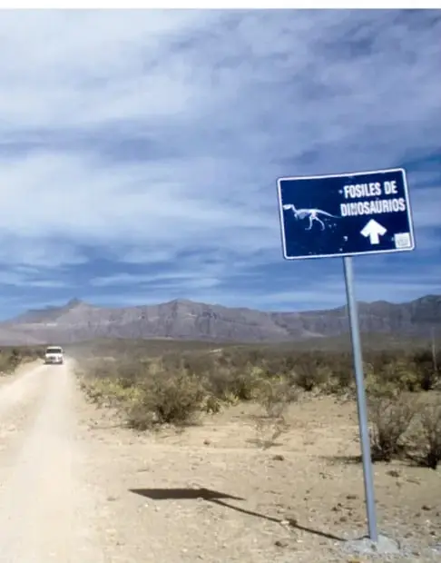 Camino de terracería en el desierto con un anuncio azul que dice «Fósiles de dinosaurios» con una flecha blanca; al fondo, montañas y un vehículo. Crédito: Salatiel Barragán Santos, Veracruz.