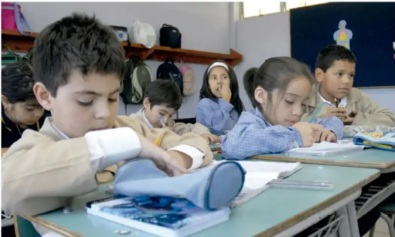 Fotografía de un salón de clases con estudiantes de primaria sentados en sus pupitres, escribiendo y leyendo. Al fondo se observa un librero con mochilas y un cartel con la silueta de un cuerpo humano.