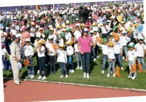 Fotografía de una cancha escolar con muchos niños y niñas uniformados formados en filas, acompañados por adultos; al frente una persona con ropa deportiva rosa dirige la actividad.