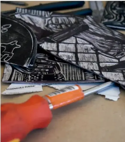 Fotografía de herramientas de artes gráficas: una matriz grabada en negro y blanco, hojas de papel y un destornillador de mango naranja sobre una mesa de trabajo.