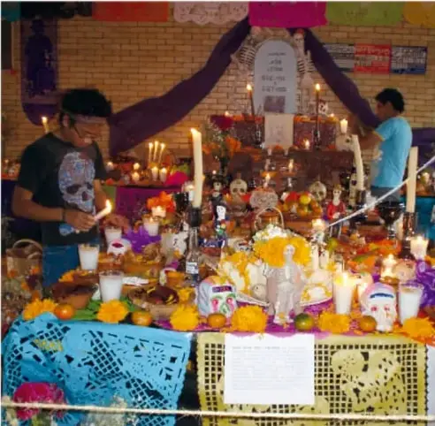 Fotografía de personas arreglando una ofrenda de Día de Muertos con velas, flores de cempasúchil, calaveritas y frutas; una tapete de papel picado azul y amarillo cuelga al frente.