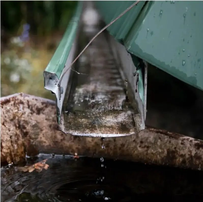 Fotografía de una canaleta o canalón metálico verde que evacúa agua hacia un recipiente de piedra, con gotas cayendo; ilustra un sistema de captación de agua de lluvia vinculado al proyecto escolar.