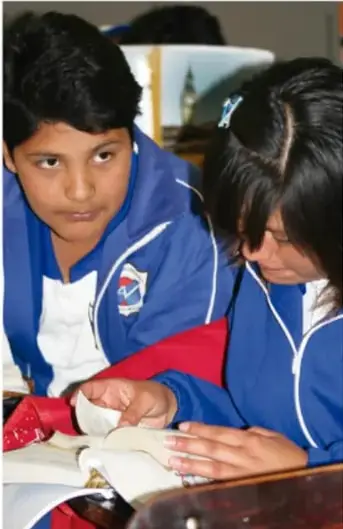 Dos estudiantes de secundaria en uniforme azul trabajando con materiales y libros sobre su escritorio.