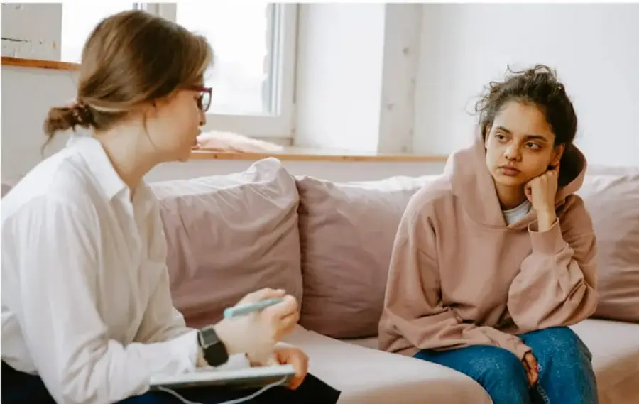 Fotografía de una sesión de acompañamiento: una profesional de la salud mental (mujer con lentes y cuaderno) conversa con una adolescente que está sentada en un sofá, con expresión seria y apoyada en la mano, en una sala luminosa.