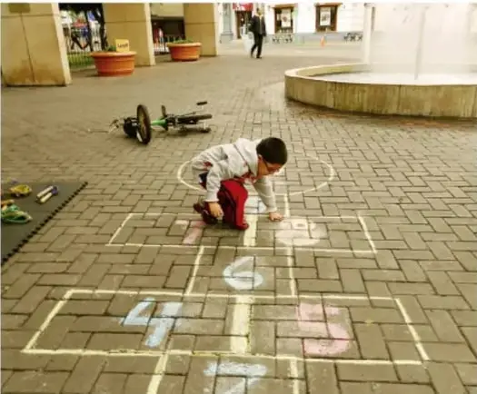 Niño agachado dibujando con gis un avión numerado en el suelo de una plaza.