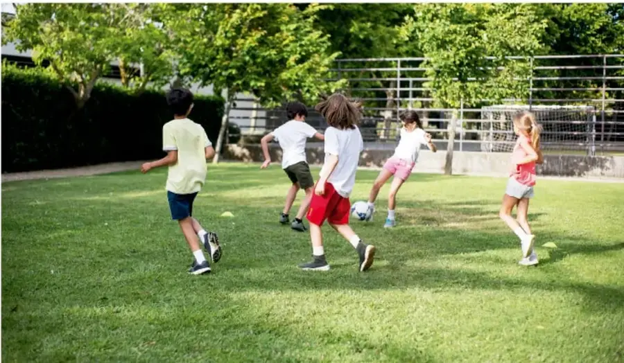 Fotografía de un grupo de niños y niñas jugando fútbol en un campo de pasto verde, con árboles y una reja al fondo. Se observa a una niña con balón y a otros corriendo.