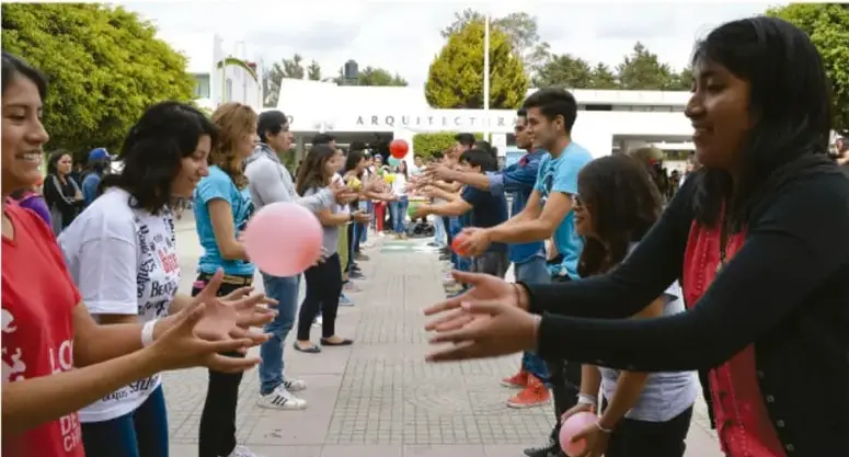 Fotografía de un grupo de jóvenes y personas adultas en un espacio al aire libre formando dos filas enfrentadas, con los brazos extendidos y globos, en una dinámica de convivencia; al fondo se observa la fachada con la leyenda 'ARQUITECTURA'. Crédito de la imagen: Guimara Schaffzin Rosales, Puebla.