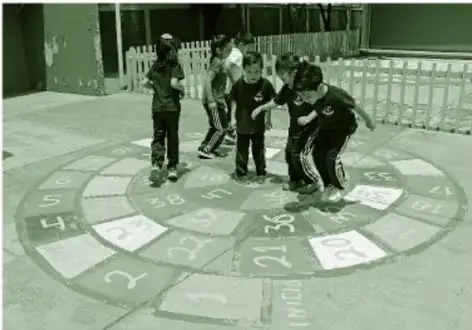 Fotografía en tono verde de niñas y niños jugando alrededor de un tablero circular pintado en el piso con casillas numeradas (estilo juego de la oca), sobre pavimento.
