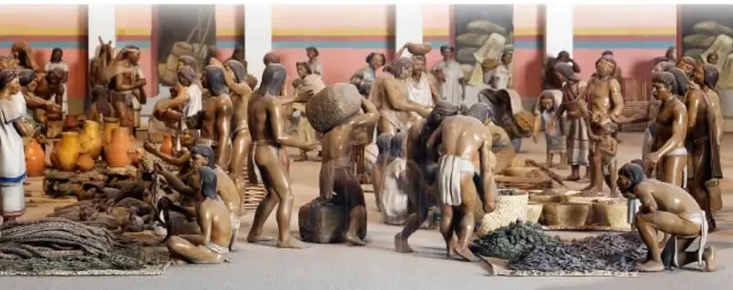 Maqueta o ilustración de un mercado mesoamericano con personas intercambiando productos como cerámica, textiles y alimentos.