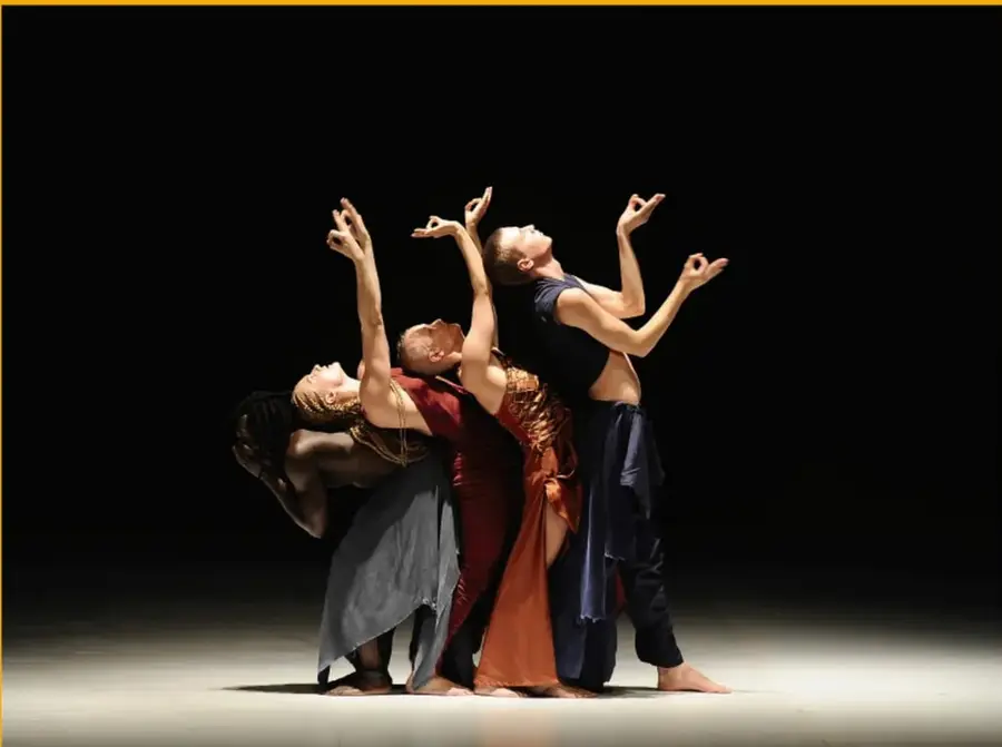 Fotografía de cuatro bailarines de la obra Aajej de Virpi Pahkinen, con vestuarios en tonos gris, rojo y azul, sobre un escenario oscuro iluminado desde el frente. Los cuerpos se entrelazan en una composición escultórica con brazos elevados.