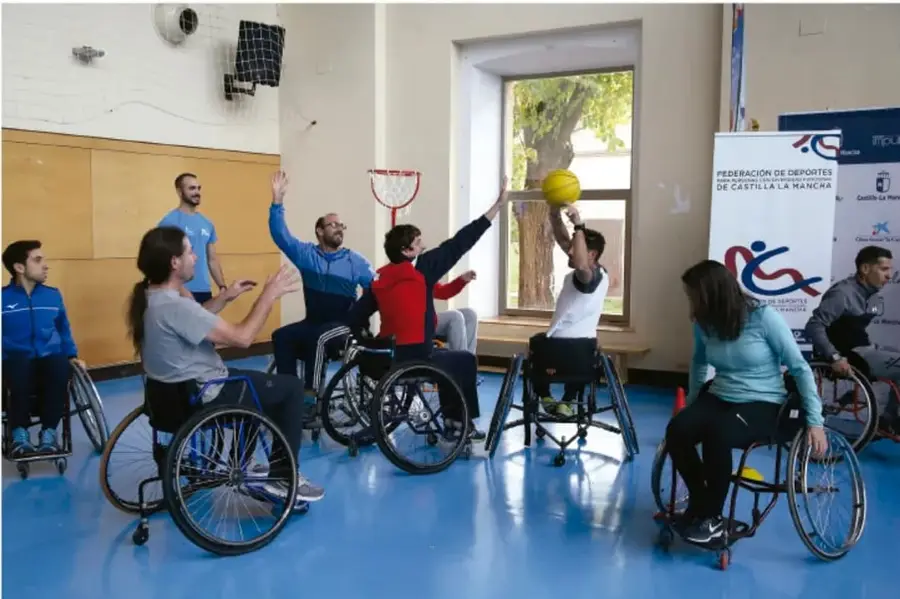 Fotografía de un grupo de personas, varias en sillas de ruedas, jugando con un balón amarillo en un patio o gimnasio escolar. Al fondo se ve un cartel de la Federación de Deportes de Castilla-La Mancha, indicando una actividad deportiva inclusiva.