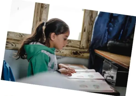 Fotografía de una niña con uniforme verde escribiendo en un cuaderno junto a una ventana de madera, dentro de un aula escolar.