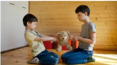 Fotografía de dos niños sentados en el piso de madera, conversando mientras juegan con un oso de peluche y cajas rojas.
