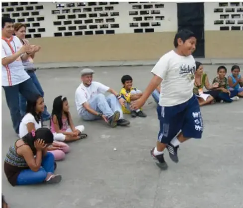 Fotografía de un grupo de estudiantes y un adulto en el patio escolar; un niño corre mientras los demás observan sentados, representando un juego en comunidad.