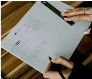 Fotografía de un plano técnico o diagrama de piso dibujado a mano sobre papel, con una regla y un bolígrafo sostenidos por manos, sobre una mesa de madera. Ilustra la representación visual ordenada de información.