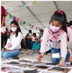 Fotografía de una niña con cubrebocas trabajando sobre un periódico mural o collage en el suelo, junto a otras personas, en una actividad escolar comunitaria.