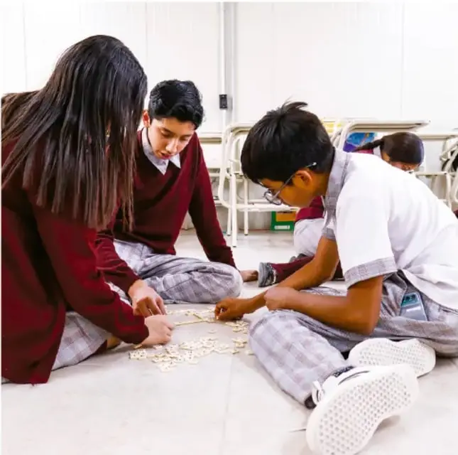 Fotografía de estudiantes de telesecundaria sentados en el piso jugando con piezas de un memorama; visten uniforme escolar con chamarra vino y pantalón a cuadros.