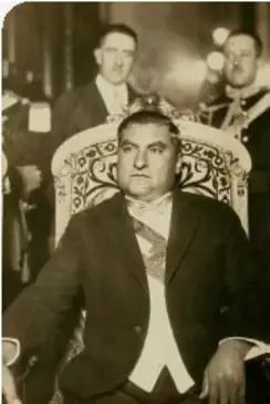 Fotografía en blanco y negro de un hombre de traje y corbata sentado, con otras personas detrás; corresponde a Emilio Portes Gil.