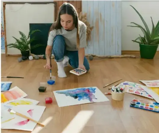 Niña arrodillada en el piso de madera pintando con acuarelas sobre varias hojas; tiene paleta, pinceles y vasos de agua alrededor, junto a plantas.