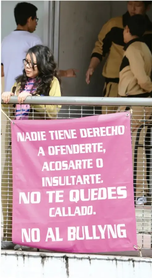 Fotografía de estudiantes en un pasillo escolar; una estudiante con lentes y suéter amarillo sostiene una pancarta rosa con el mensaje: 'NADIE TIENE DERECHO A OFENDERTE, ACOSARTE O INSULTARTE. NO TE QUEDES CALLADO. NO AL BULLYNG'.