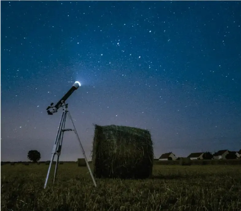 Fotografía de un telescopio apuntando al cielo estrellado en un campo abierto, con un fardo de heno y casas al fondo.