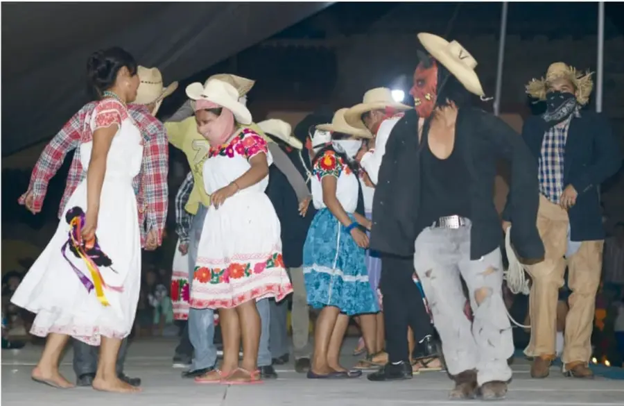 Fotografía de una danza del Xantolo en Veracruz: varios danzantes con sombreros de paja, un hombre enmascarado de diablo con camisa negra y pantalones rotos, y mujeres con vestidos blancos bordados, descalzas, bailando en un escenario.