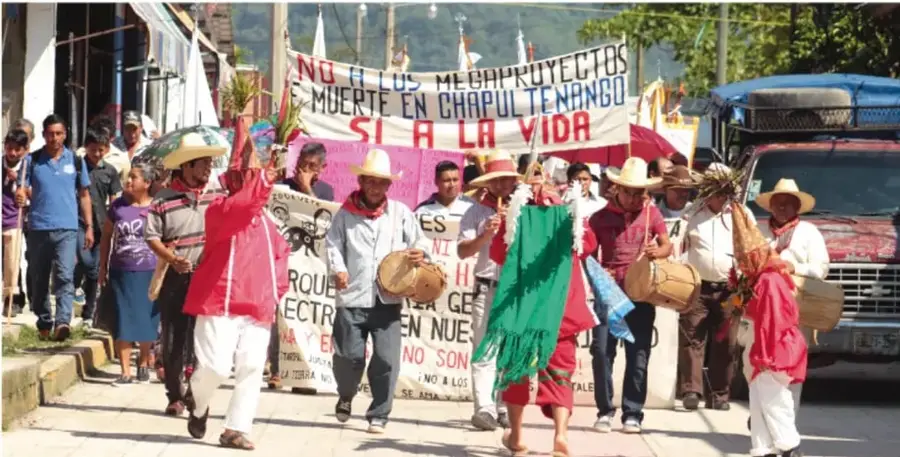 Fotografía de una marcha indígena en Chiapas. Los manifestantes portan pancartas con leyendas como 'NO A LOS MEGAPROYECTOS', 'MUERTE EN CHAPULTENANGO', 'SÍ A LA VIDA'. Visten ropas tradicionales y algunos llevan sombreros. Al fondo se observa una camioneta y edificios.