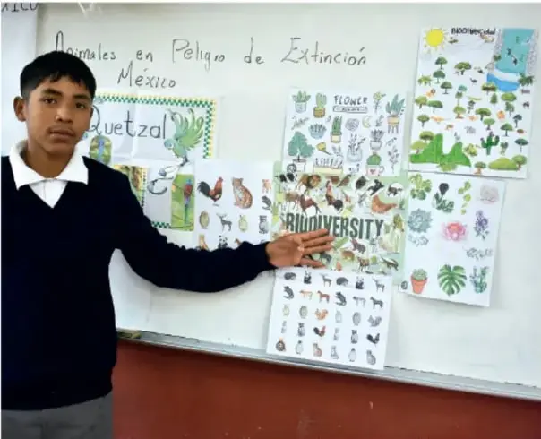 Fotografía de un estudiante de Telesecundaria frente a un pizarrón con carteles sobre 'Animales en Peligro de Extinción en México', 'Quetzal', 'BIODIVERSITY' y 'FLOWER', mostrando sus trabajos.