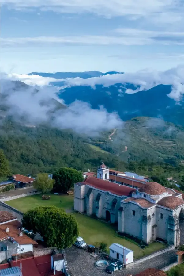Fotografía aérea de un templo colonial de piedra con cúpula y campanario, rodeado de jardines, casas de tejado rojo y montañas cubiertas de nubes y bosque.