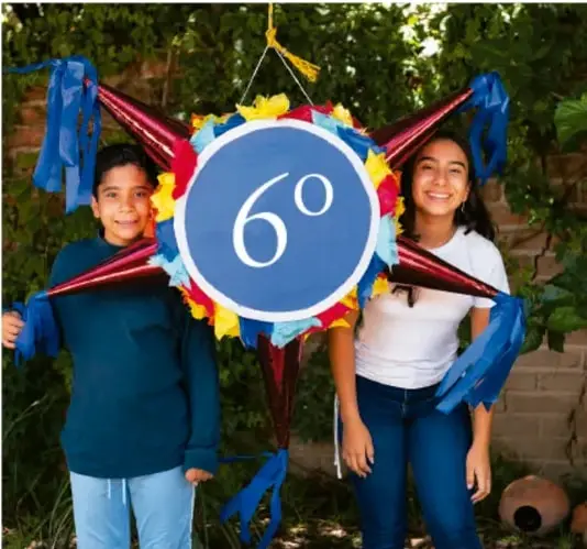 Fotografía de dos estudiantes (niño y niña) sosteniendo una piñata con el número 6° en un círculo azul, rodeada de listones de colores.