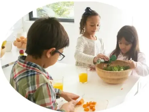 Fotografía de tres niñas y niños preparando una ensalada en la cocina: un niño con camisa a cuadros y lentes corta zanahorias al frente, mientras dos niñas agregan verduras a un tazón de madera con jugo de naranja en la mesa.