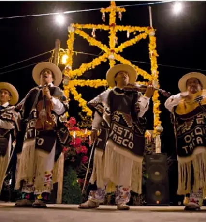 Fotografía de músicos p'urhépecha con sombreros y trajes tradicionales interpretando pirekuas en una celebración nocturna con un arco floreado iluminado. Crédito: Francisco Palma Lagunas, Ciudad de México.