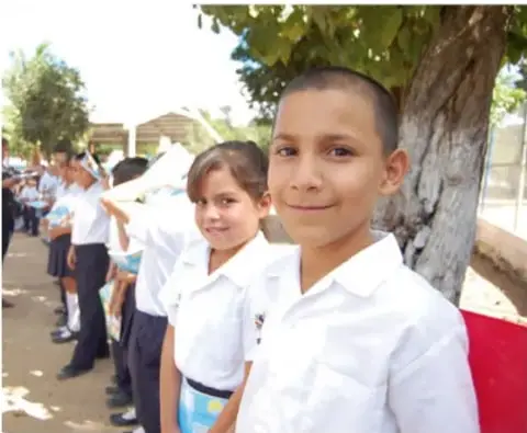 Fotografía de estudiantes de primaria uniformados, formados en el patio de la escuela, con árboles y edificios al fondo.