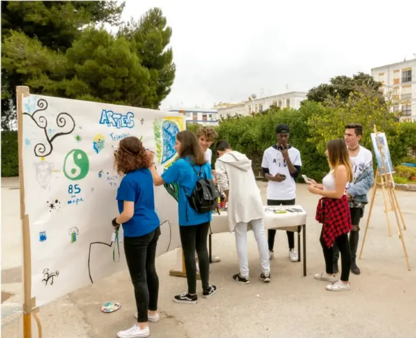 Fotografía de un grupo de jóvenes estudiantes pintando y preparando un mural o manta sobre un armazón de madera en un patio exterior, al aire libre, con edificios y árboles al fondo; representan el trabajo colaborativo del proyecto.
