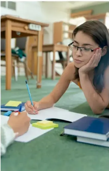 Fotografía de una estudiante con lentes escribiendo en su cuaderno, con libros y un teléfono sobre el escritorio, en una biblioteca o salón de estudio.