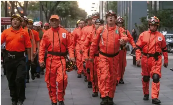 Grupo de personas con uniformes naranjas y cascos caminando por una calle, posiblemente cuerpos de seguridad o protección civil.