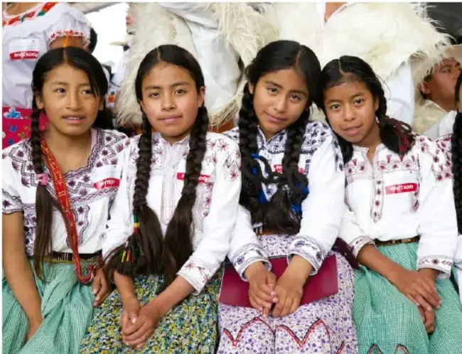 Fotografía de cuatro niñas indígenas con vestimenta tradicional bordada y trenzas, sentadas en un evento comunitario.