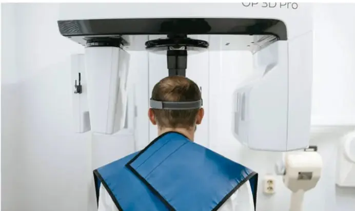 Fotografía de un equipo de rayos X panorámico dental (marca '3D PRO') con un paciente de espaldas usando protección plomada azul.