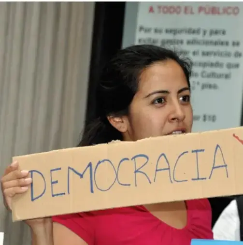 Fotografía de una mujer joven sosteniendo un cartel de cartón con la palabra DEMOCRACIA escrita a mano, en un espacio público con un letrero de fondo que dice 'A TODO EL PÚBLICO'.